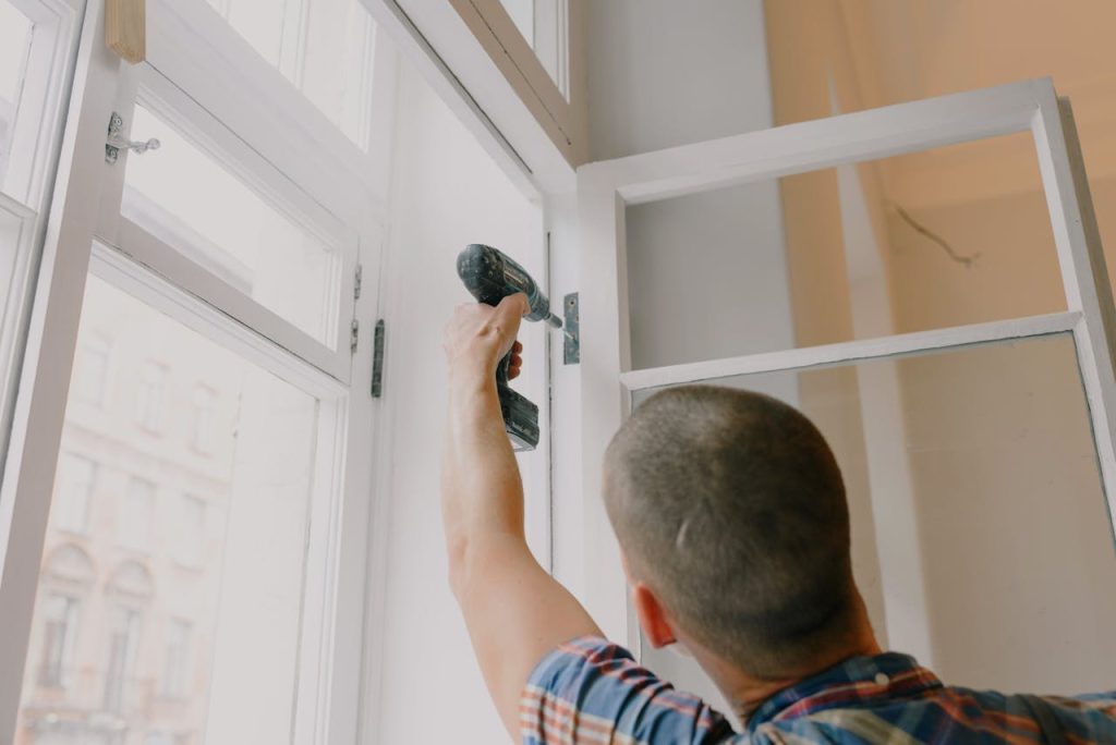 Back view of unrecognizable craftsman using cordless screwdriver and assembling window while screwing metal hinge during renovation works in apartment