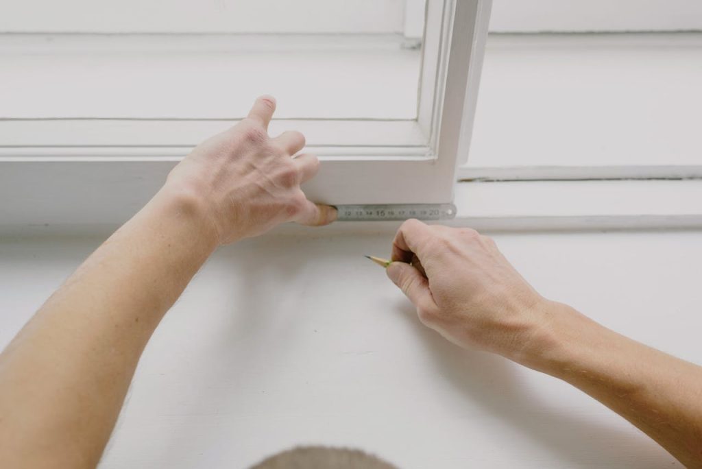 From above of crop unrecognizable male worker measuring window frame with metal ruler and pencil in house