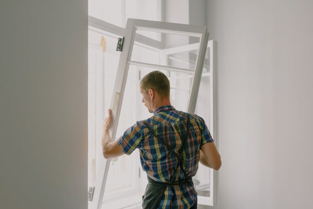 Back view of anonymous male fitter in checkered shirt and apron mounting window with hinges near white walls at home